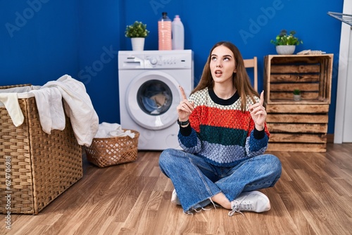Young hispanic girl doing laundry amazed and surprised looking up and pointing with fingers and raised arms.