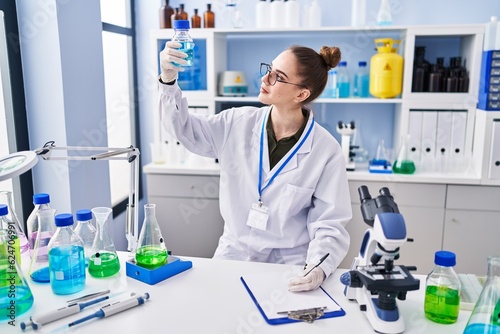 Young woman scientist writing on clipboard holding test tube at laboratory