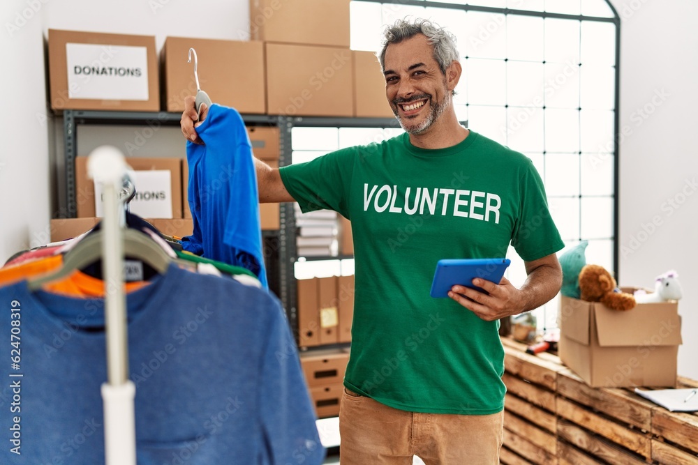 Middle age grey-haired man volunteer using touchpad holding clothes at charity center Middle age grey-haired man volunteer using touchpad holding clothes at charity center