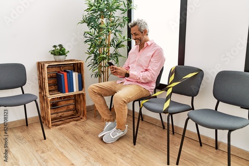 Middle age grey-haired man using smartphone sitting on chair at waiting room