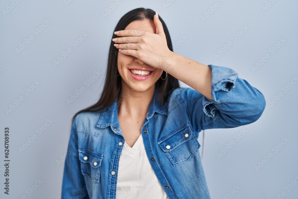 Hispanic woman standing over blue background smiling and laughing with hand on face covering eyes for surprise. blind concept. Hispanic woman standing over blue background smiling and laughing with hand on face covering eyes for surprise. blind concept.