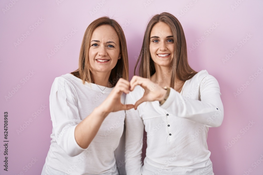 Middle age mother and young daughter standing over pink background smiling in love doing heart symbol shape with hands. romantic concept. Middle age mother and young daughter standing over pink background smiling in love doing heart symbol shape with hands. romantic concept.