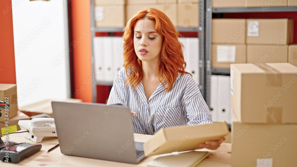 Young redhead woman ecommerce business worker using laptop holding package at office Young redhead woman ecommerce business worker using laptop holding package at office