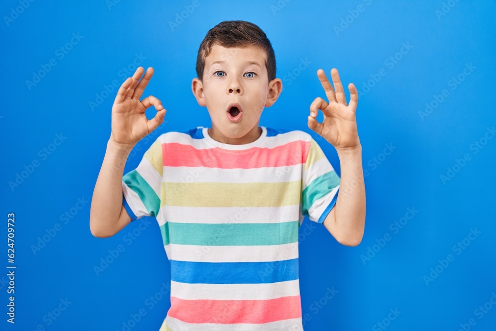 Young caucasian kid standing over blue background looking surprised and shocked doing ok approval symbol with fingers. crazy expression Young caucasian kid standing over blue background looking surprised and shocked doing ok approval symbol with fingers. crazy expression