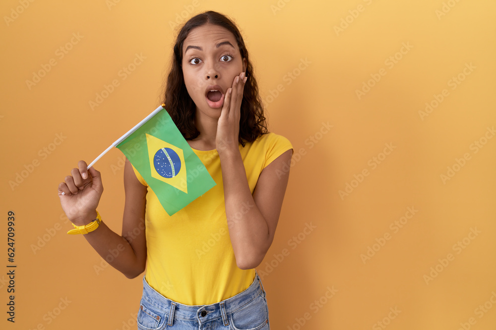 Young hispanic woman holding brazil flag afraid and shocked, surprise and amazed expression with hands on face Young hispanic woman holding brazil flag afraid and shocked, surprise and amazed expression with hands on face