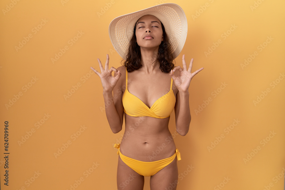 Young hispanic woman wearing bikini and summer hat relaxed and smiling with eyes closed doing meditation gesture with fingers. yoga concept. Young hispanic woman wearing bikini and summer hat relaxed and smiling with eyes closed doing meditation gesture with fingers. yoga concept.