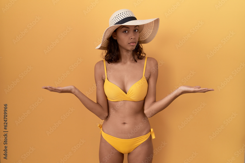 Young hispanic woman wearing bikini and summer hat clueless and confused with open arms, no idea concept. Young hispanic woman wearing bikini and summer hat clueless and confused with open arms, no idea concept.