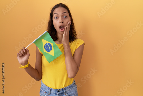 Young hispanic woman holding brazil flag afraid and shocked  surprise and amazed expression with hands on face