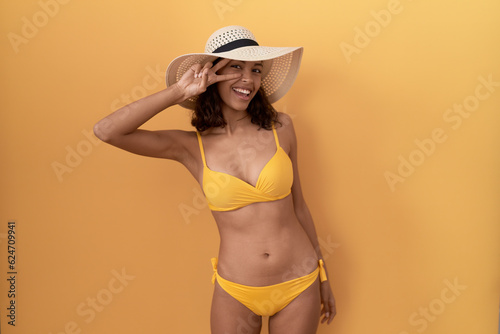Young hispanic woman wearing bikini and summer hat doing peace symbol with fingers over face  smiling cheerful showing victory