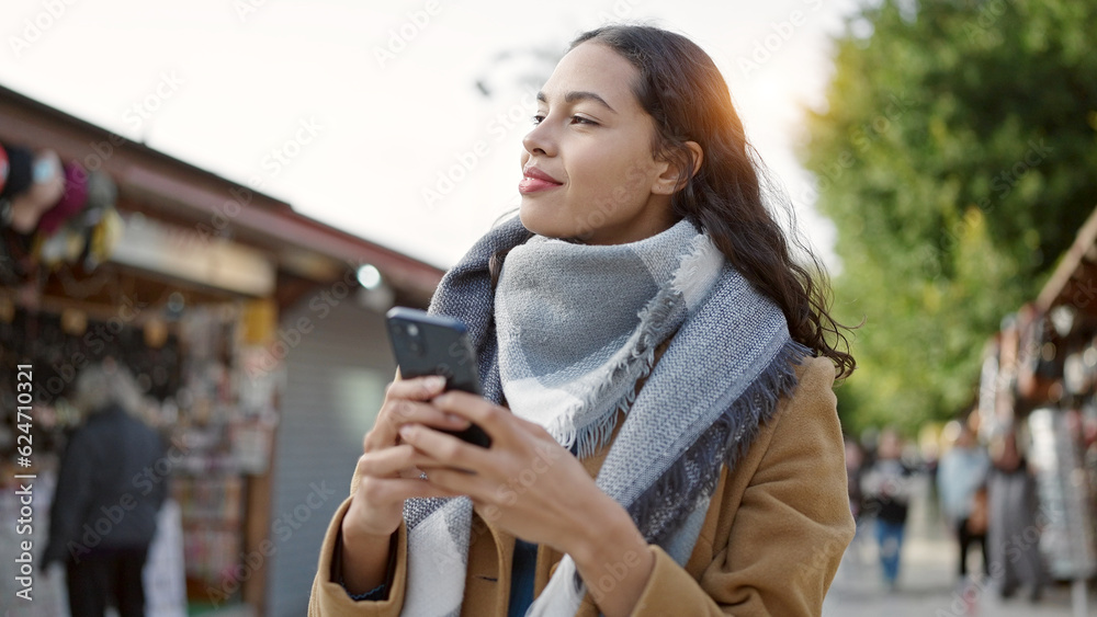 Fototapeta premium Young beautiful hispanic woman using smartphone smiling at street market