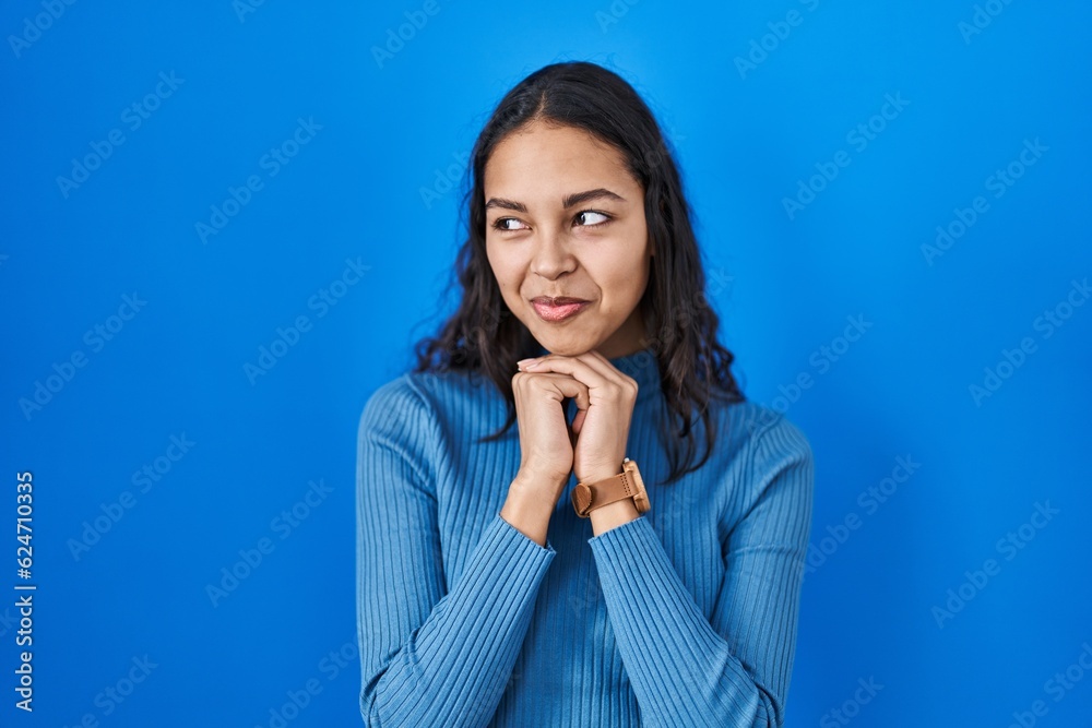 Young brazilian woman standing over blue isolated background laughing nervous and excited with hands on chin looking to the side Young brazilian woman standing over blue isolated background laughing nervous and excited with hands on chin looking to the side