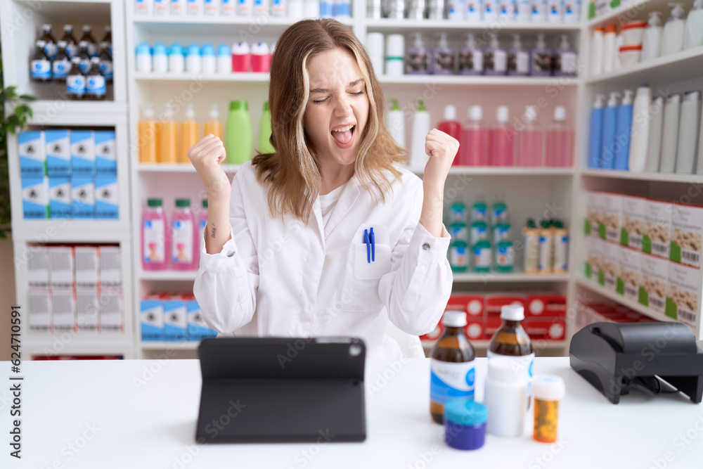 Young caucasian woman working at pharmacy drugstore using tablet very happy and excited doing winner gesture with arms raised, smiling and screaming for success. celebration concept.