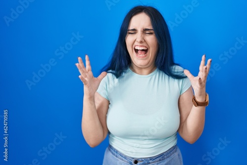 Young modern girl with blue hair standing over blue background celebrating mad and crazy for success with arms raised and closed eyes screaming excited. winner concept