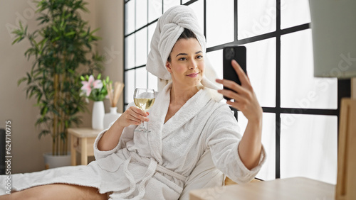 Young beautiful hispanic woman wearing bathrobe drinking champagne make selfie by smartphone at bedroom