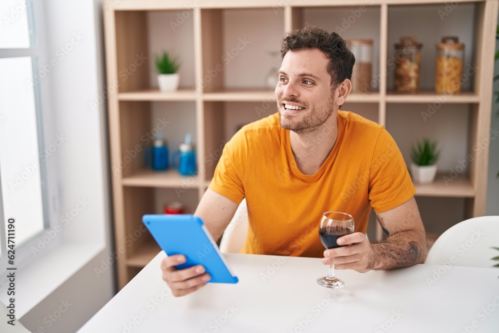 Fototapeta premium Young hispanic man using touchpad drinkng wine at home