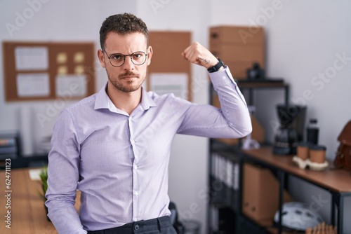 Young hispanic man at the office strong person showing arm muscle, confident and proud of power