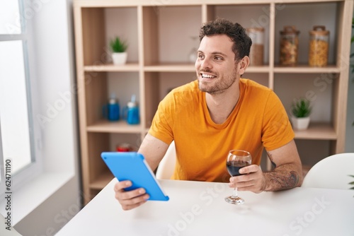 Young hispanic man using touchpad drinkng wine at home