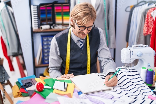 Young blond man tailor smiling confident drawing on notebook at clothing factory