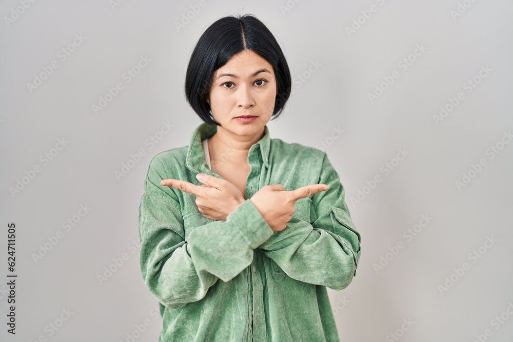 Young asian woman standing over white background pointing to both sides with fingers, different direction disagree