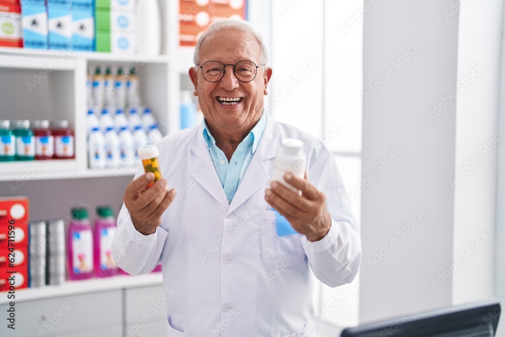Senior grey-haired man pharmacist smiling confident holding pills bottles at pharmacy
