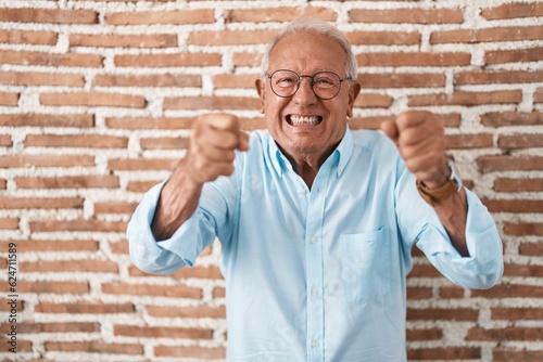 Senior man with grey hair standing over bricks wall angry and mad raising fists frustrated and furious while shouting with anger. rage and aggressive concept.