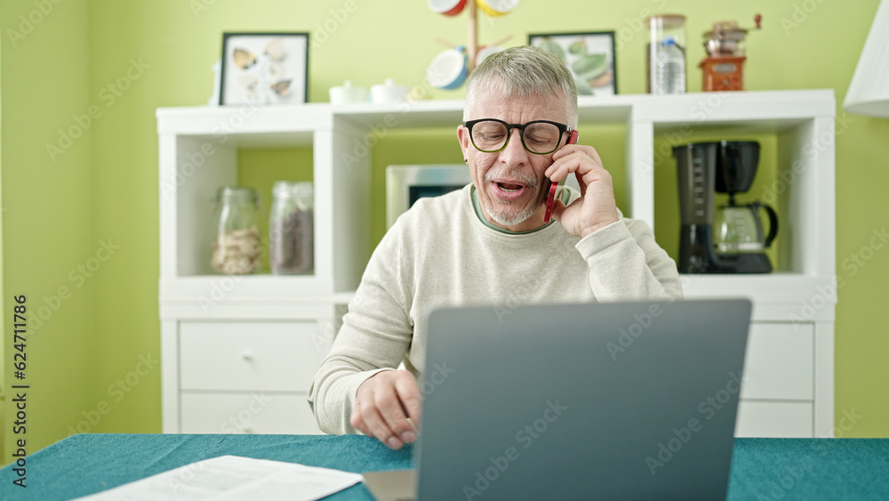 Middle age grey-haired man talking on smartphone using laptop at home Middle age grey-haired man talking on smartphone using laptop at home