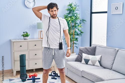 Handsome latin man wearing sportswear at home confuse and wonder about question. uncertain with doubt, thinking with hand on head. pensive concept. photo