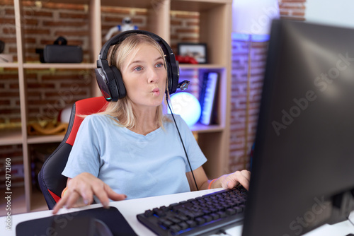 Young caucasian woman playing video games wearing headphones making fish face with lips, crazy and comical gesture. funny expression.