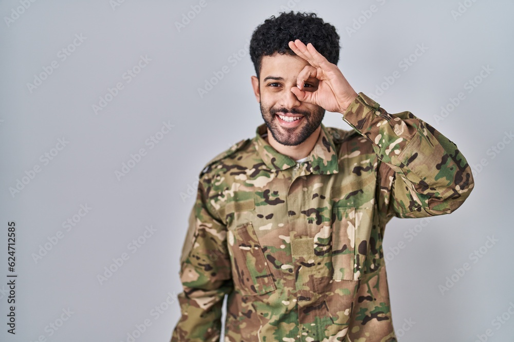 Arab man wearing camouflage army uniform doing ok gesture with hand smiling, eye looking through fingers with happy face.
