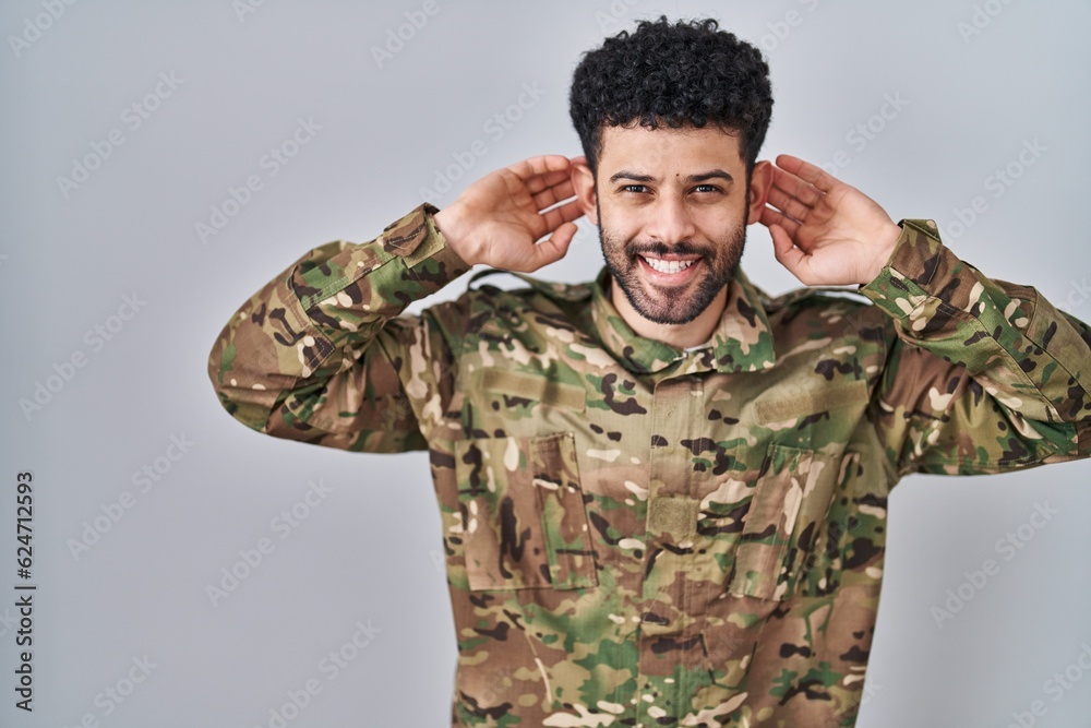 Arab man wearing camouflage army uniform smiling pulling ears with fingers, funny gesture. audition problem