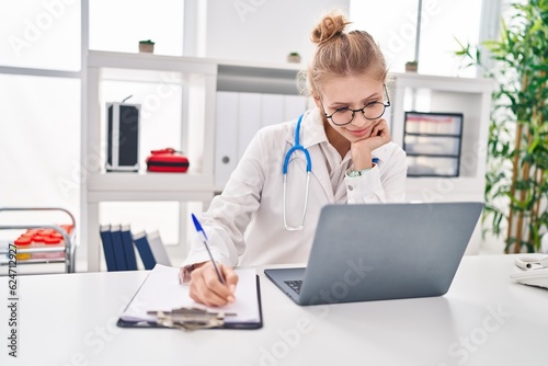 Young blonde woman doctor using laptop writing medical report at clinic