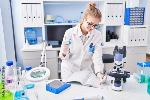 Young blonde woman scientist writing on notebook holding test tube at laboratory