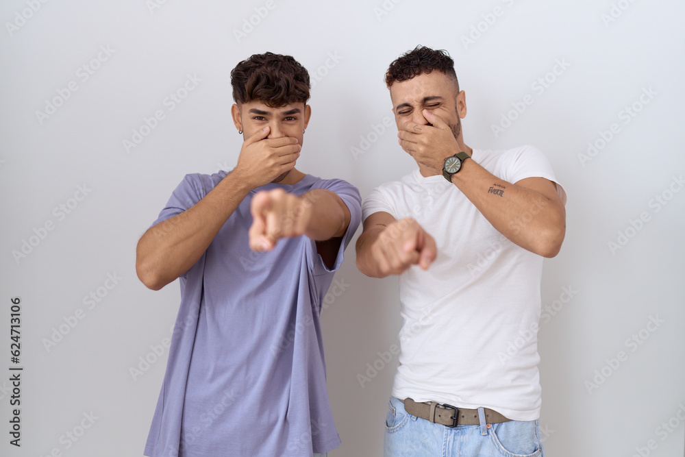 Homosexual gay couple standing over white background laughing at you, pointing finger to the camera with hand over mouth, shame expression Homosexual gay couple standing over white background laughing at you, pointing finger to the camera with hand over mouth, shame expression