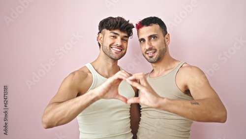 Two men couple hugging each other doing heart gesture over isolated pink background