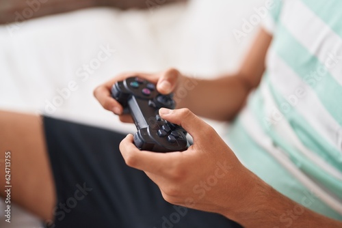 Young hispanic teenager playing video game sitting on bed at bedroom