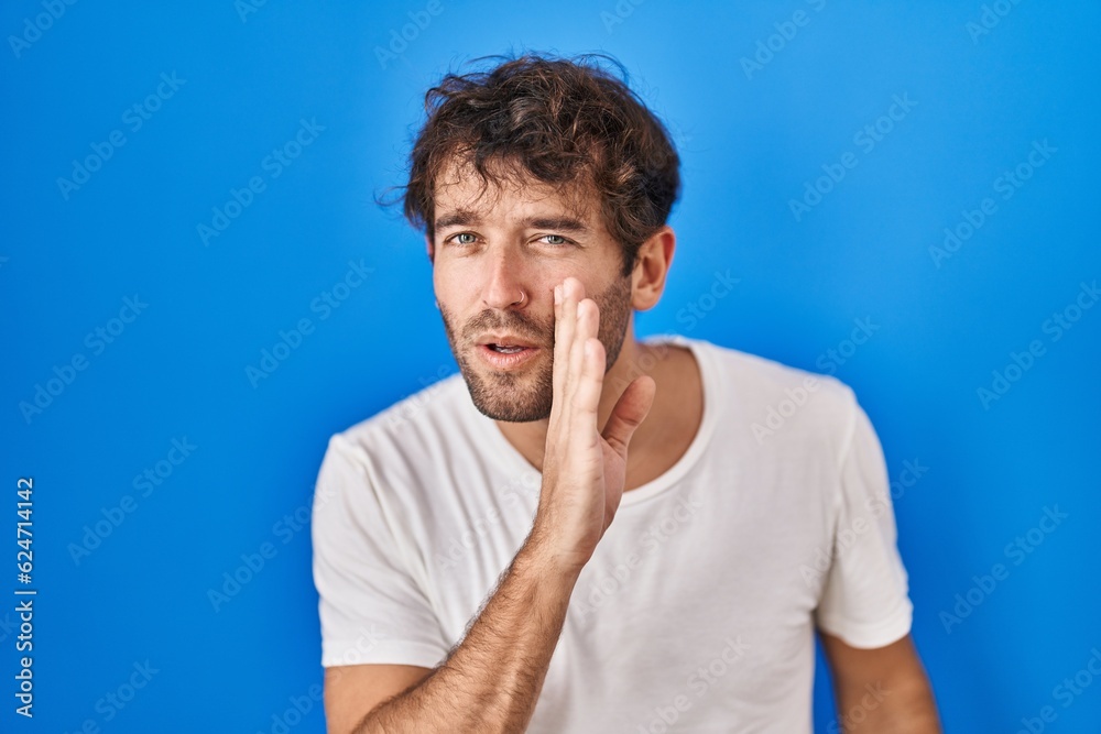 Hispanic young man standing over blue background hand on mouth telling secret rumor, whispering malicious talk conversation Hispanic young man standing over blue background hand on mouth telling secret rumor, whispering malicious talk conversation