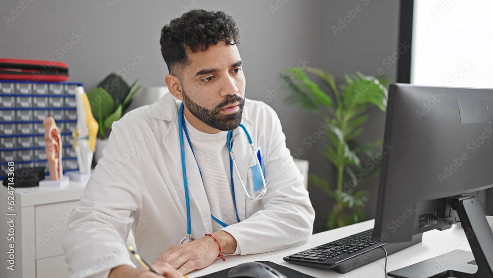 Young hispanic man doctor using computer writing notes at clinic Young hispanic man doctor using computer writing notes at clinic