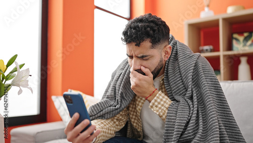 Foto Young hispanic man sitting on sofa having online medical consultation coughing a