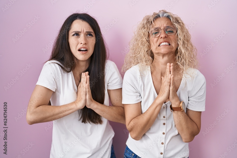 Mother and daughter standing together over pink background begging and praying with hands together with hope expression on face very emotional and worried. begging. Mother and daughter standing together over pink background begging and praying with hands together with hope expression on face very emotional and worried. begging.