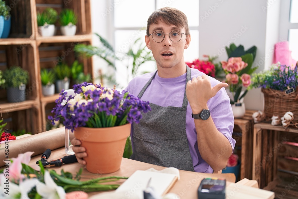 Caucasian blond man working at florist shop surprised pointing with hand finger to the side, open mouth amazed expression.