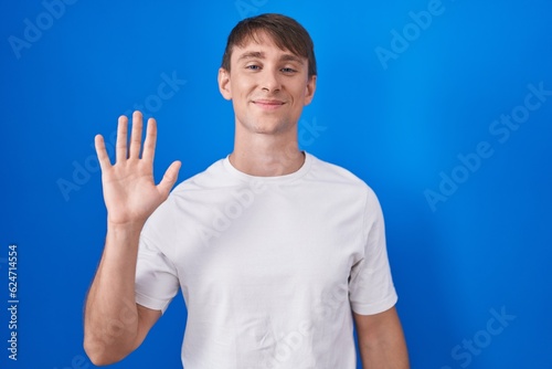 Caucasian blond man standing over blue background waiving saying hello happy and smiling, friendly welcome gesture