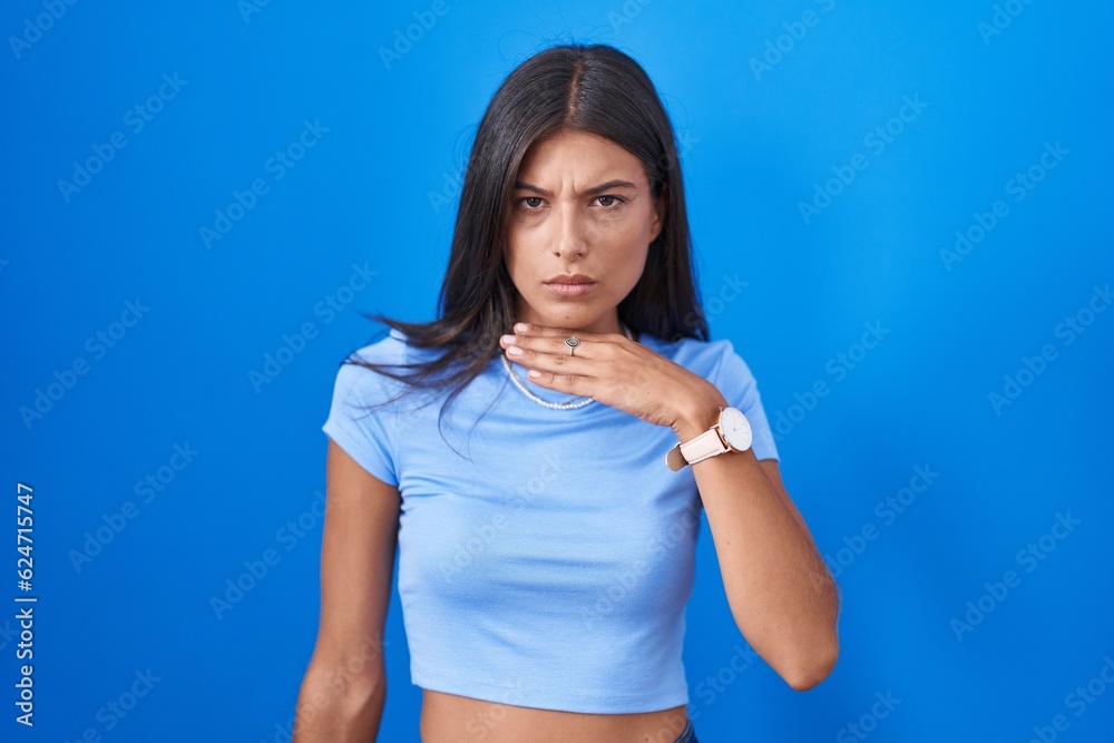 Brunette young woman standing over blue background cutting throat with hand as knife, threaten aggression with furious violence
