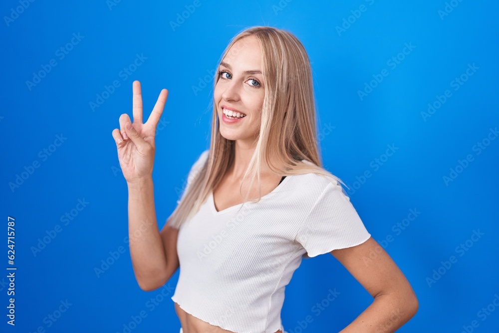 Fototapeta premium Young caucasian woman standing over blue background smiling looking to the camera showing fingers doing victory sign. number two.