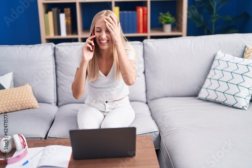 Young blonde woman talking on smartphone using laptop at home