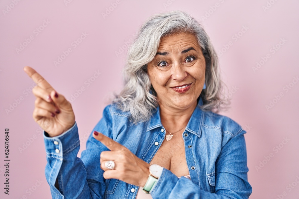 Obraz premium Middle age woman with grey hair standing over pink background smiling and looking at the camera pointing with two hands and fingers to the side.