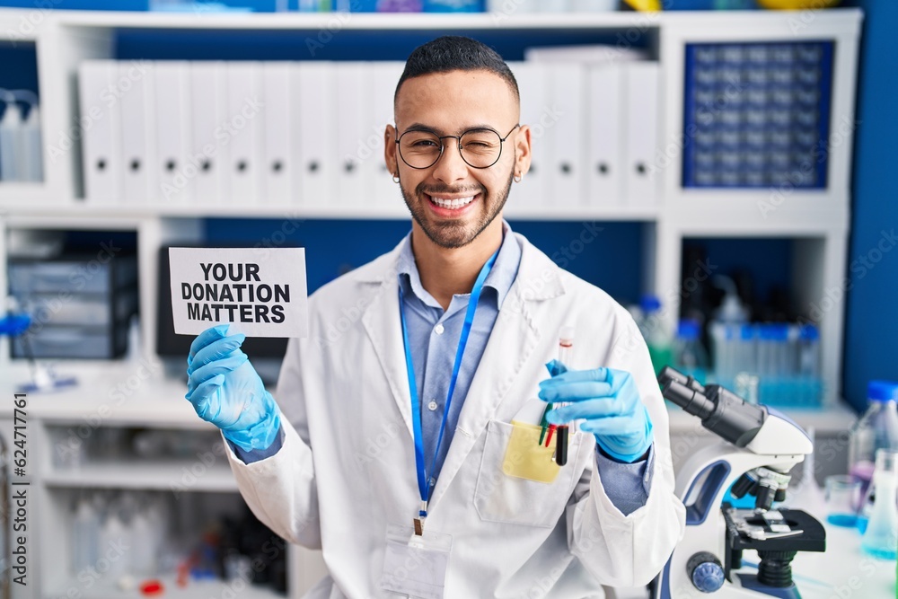 Young hispanic man working at scientist laboratory holding your donation matters holding blood sample winking looking at the camera with sexy expression, cheerful and happy face. Young hispanic man working at scientist laboratory holding your donation matters holding blood sample winking looking at the camera with sexy expression, cheerful and happy face.