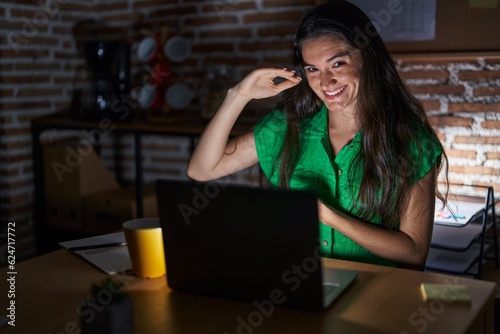 Young teenager girl working at the office at night gesturing with hands showing big and large size sign, measure symbol. smiling looking at the camera. measuring concept.