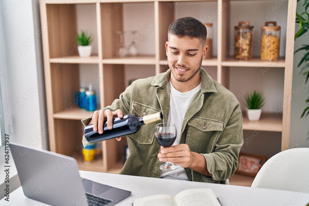 Young hispanic man using laptop pouring wine on glass at home