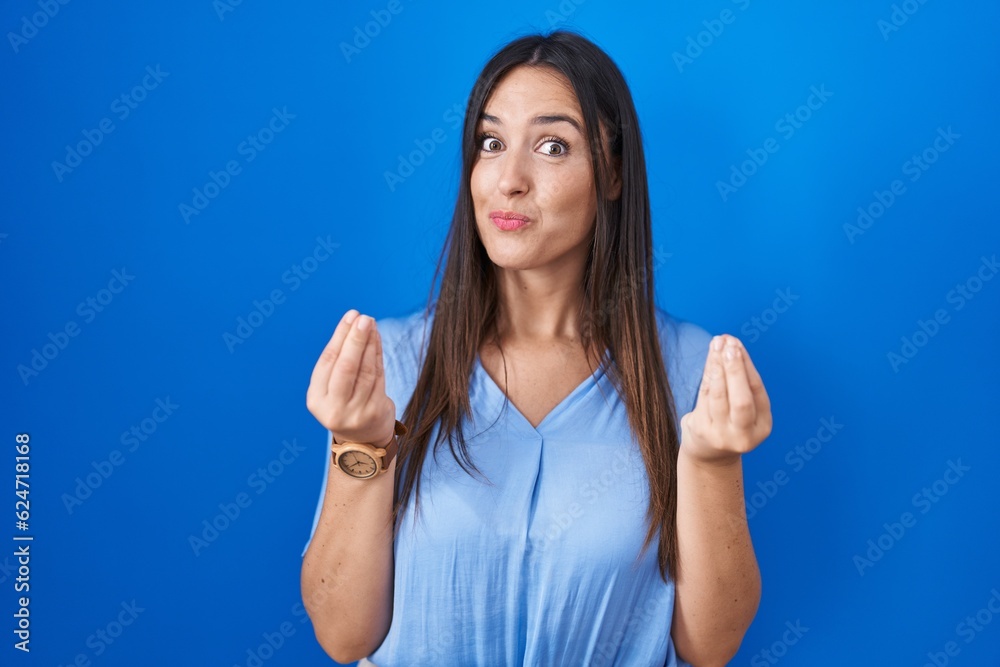 Fototapeta premium Young brunette woman standing over blue background doing money gesture with hands, asking for salary payment, millionaire business