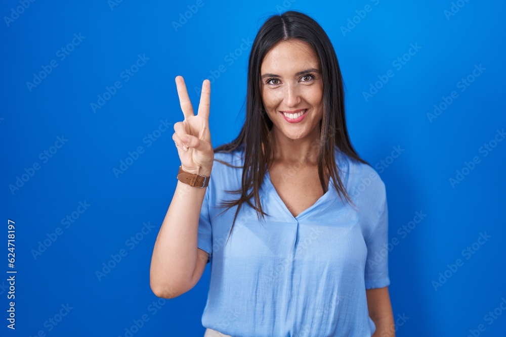 Young brunette woman standing over blue background showing and pointing up with fingers number two while smiling confident and happy. Young brunette woman standing over blue background showing and pointing up with fingers number two while smiling confident and happy.